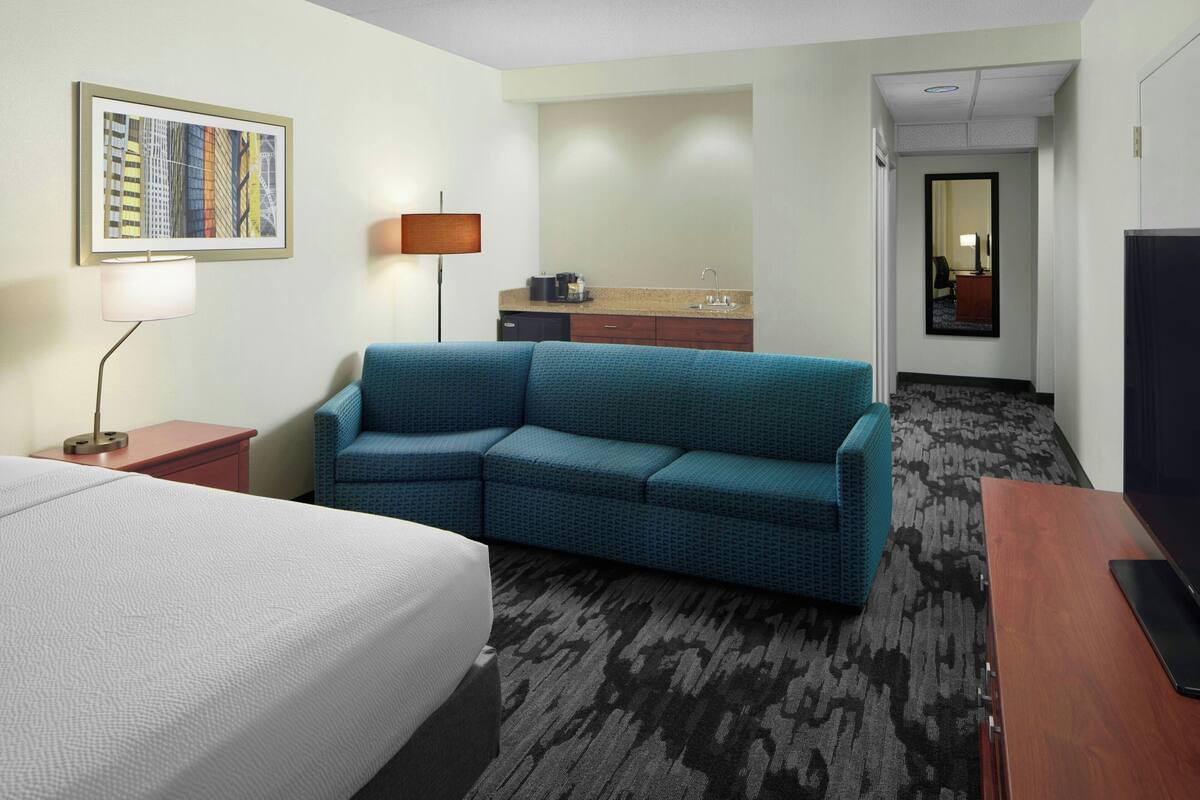 A hotel room with a bed, a sofa, a desk, a lamp, a sink, and a mirror.