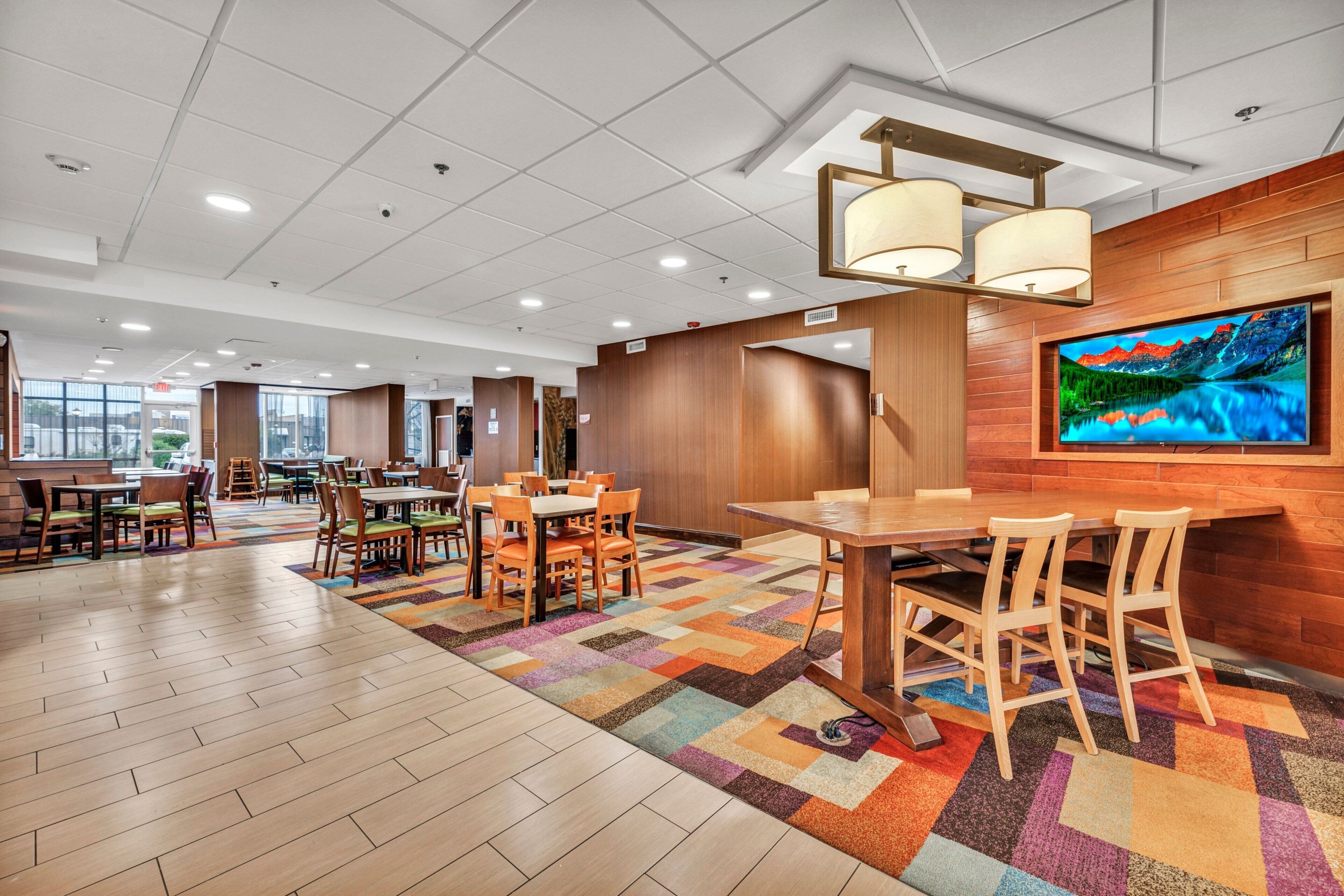 A spacious dining area with multiple tables and chairs, a colorful carpet, and a large flat-screen TV mounted on the wall.