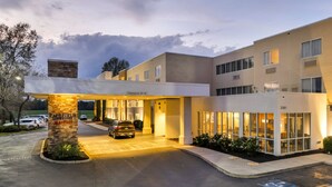Entrada de hotel con un camino cubierto, un pilar de piedra con el nombre y un área de estacionamiento.