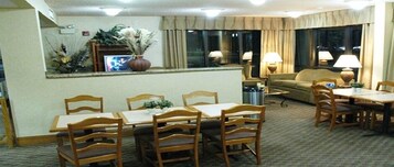 Uma área de refeições de hotel com uma mesa comprida e cadeiras, um sofá e uma tela de televisão.