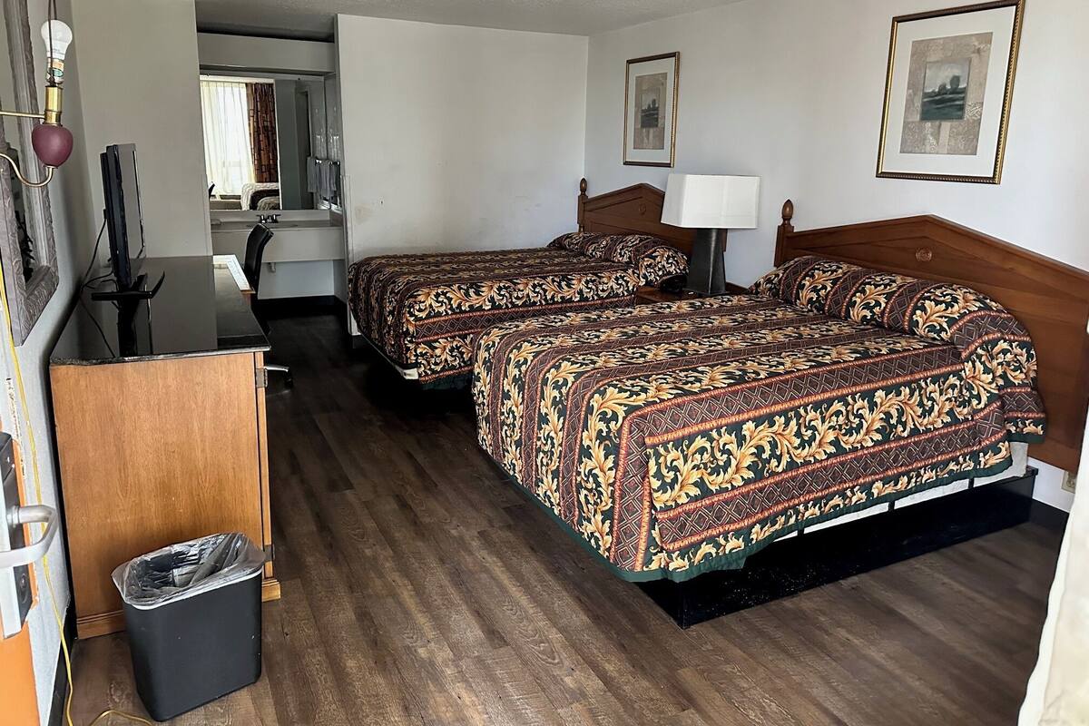 A hotel room with two beds, a desk, a chair, a trash bin, and framed pictures on the walls.