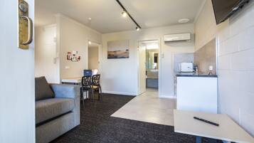 A modern living room with a grey sofa, a dining area with chairs, a kitchen with a fridge, and an air conditioning unit on the wall.