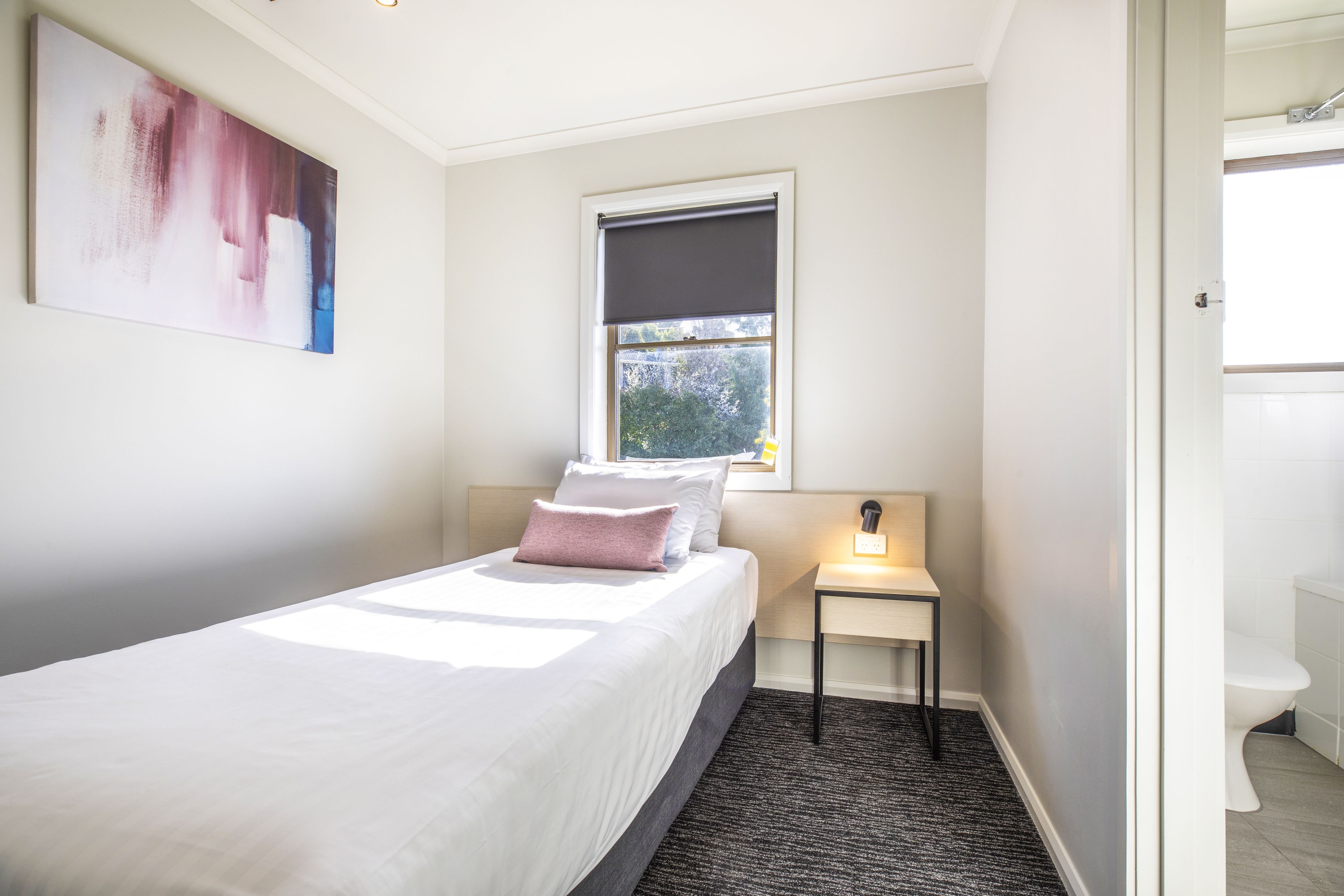 A neatly arranged bedroom with a bed, a bedside table with a lamp, a window with a view, and a bathroom visible through an open door.