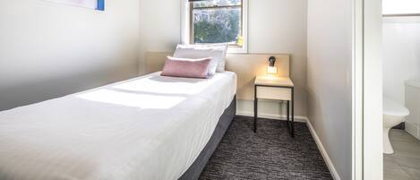A neatly arranged bedroom with a bed, a bedside table with a lamp, a window with a view, and a bathroom visible through an open door.