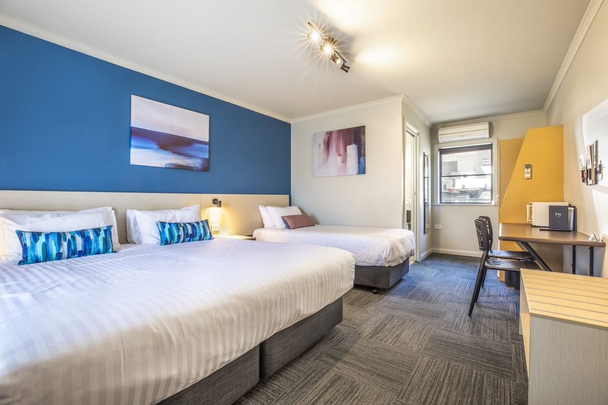 A hotel room with two beds, a desk, and a chair. There are paintings on the walls and a window providing natural light.