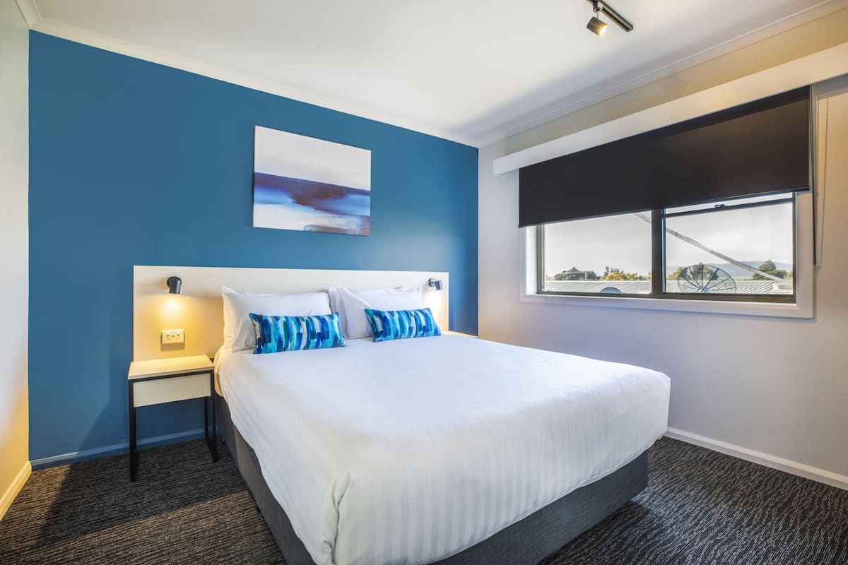 A hotel room with a large bed, a nightstand, a wall-mounted painting, and a window with a view of a bridge and landscape.