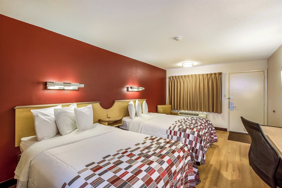 Habitación de hotel con dos camas, un escritorio, una silla, una ventana con cortinas y una puerta.