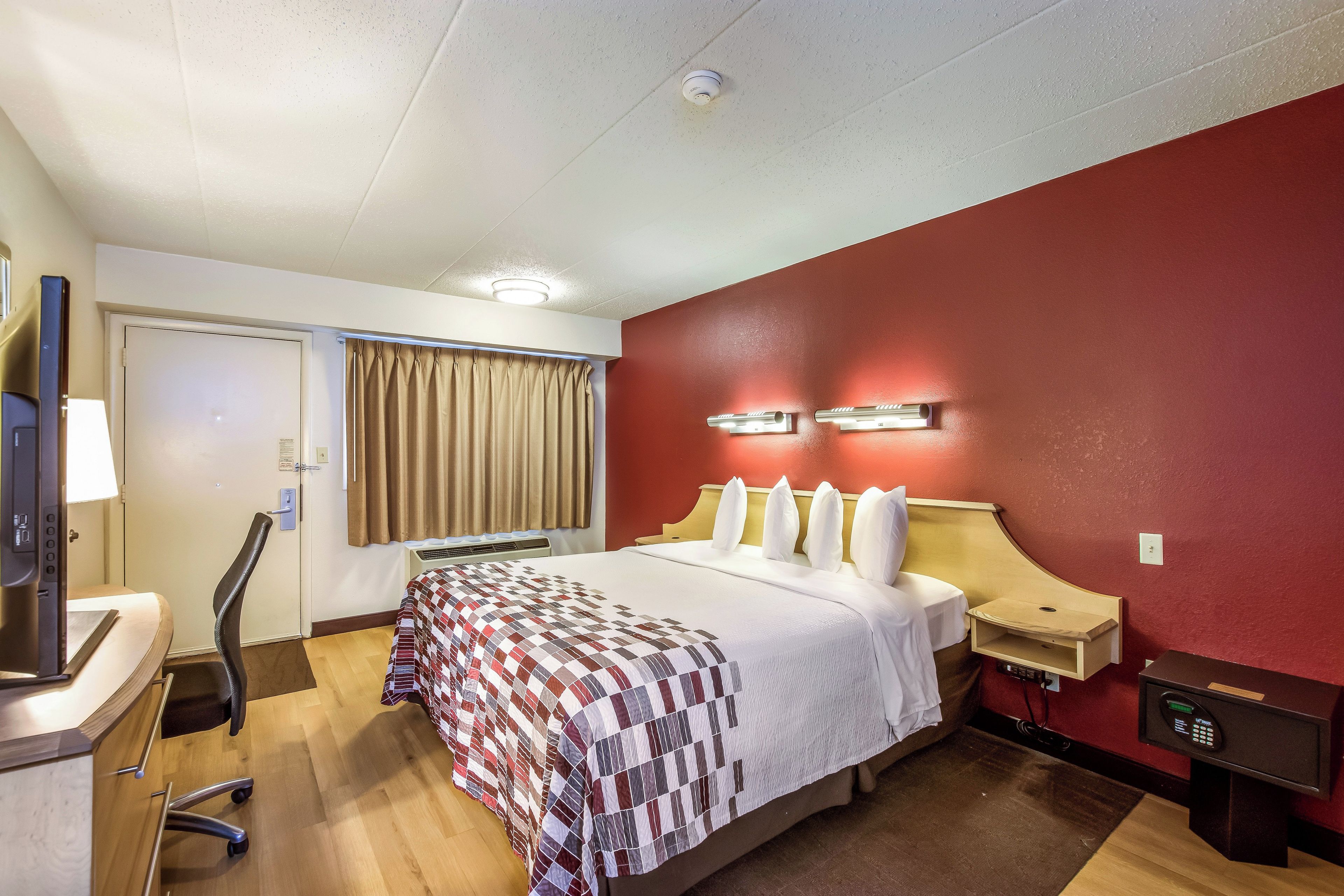 Une chambre d’hôtel avec un lit, un bureau, une chaise, une télévision, un mur rouge et un luminaire au plafond.