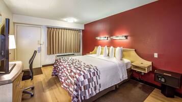 Une chambre d’hôtel avec un lit, un bureau, une chaise, une télévision, un mur rouge et un luminaire au plafond.