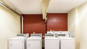 A laundry room with two front-loading washing machines and a dryer, white walls, and a red accent wall. - Red Roof Inn Detroit - Plymouth/ Canton (Plymouth)