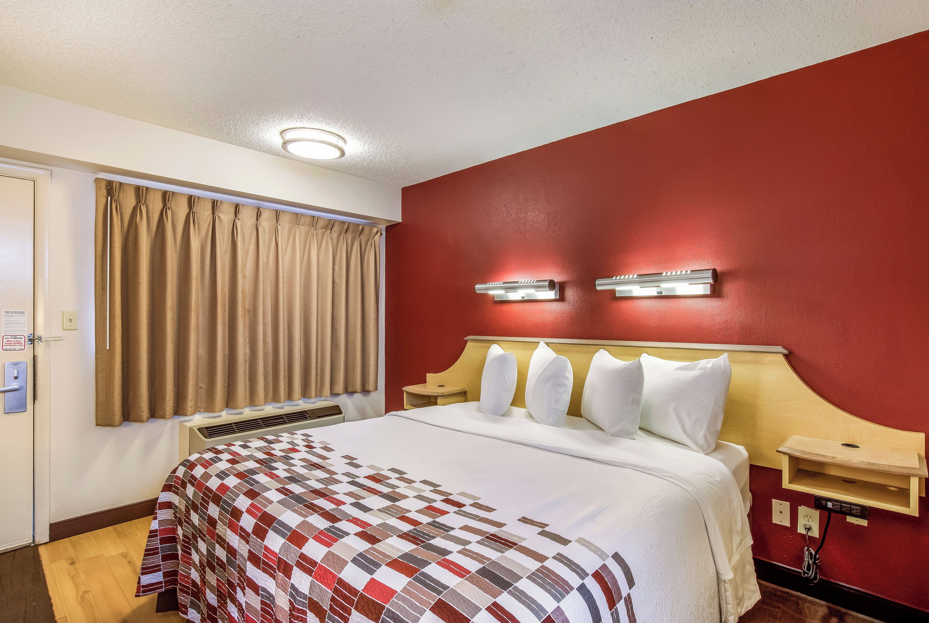 A hotel room with a bed, curtains, a wall-mounted light, and a bedside table.
