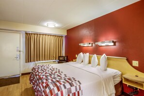 A hotel room with a bed, a TV, a bedside table, and a red accent wall. - Red Roof Inn Detroit - Plymouth/ Canton (Plymouth)