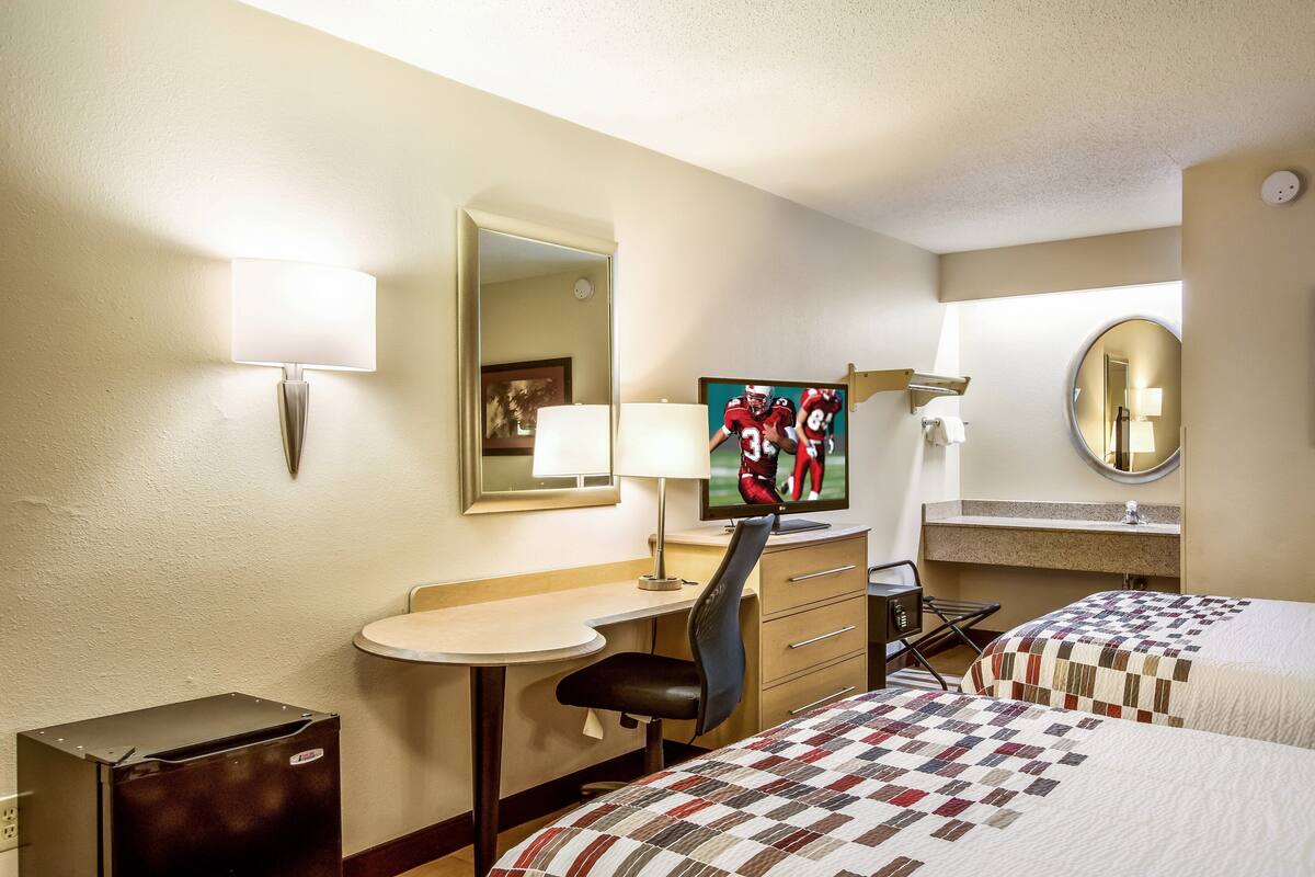 A hotel room with two beds, a desk, a chair, a television, a mirror, and a small trash bin.