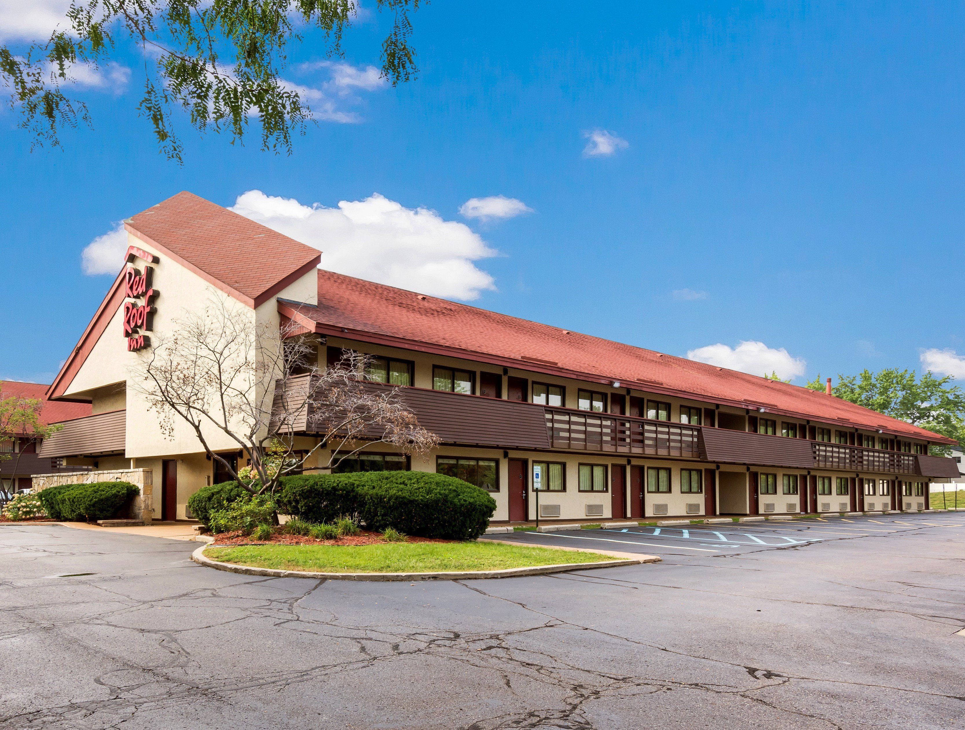 A motel with a red roof and a sign that reads 'Red Roof Inn.' It has a parking lot in front with a few trees and shrubs.