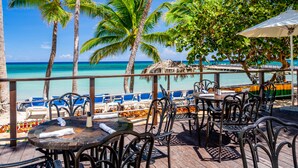 Eine Terrasse mit Tischen und Stühlen mit Blick auf einen Strand, Palmen und einen klaren, blauen Himmel.