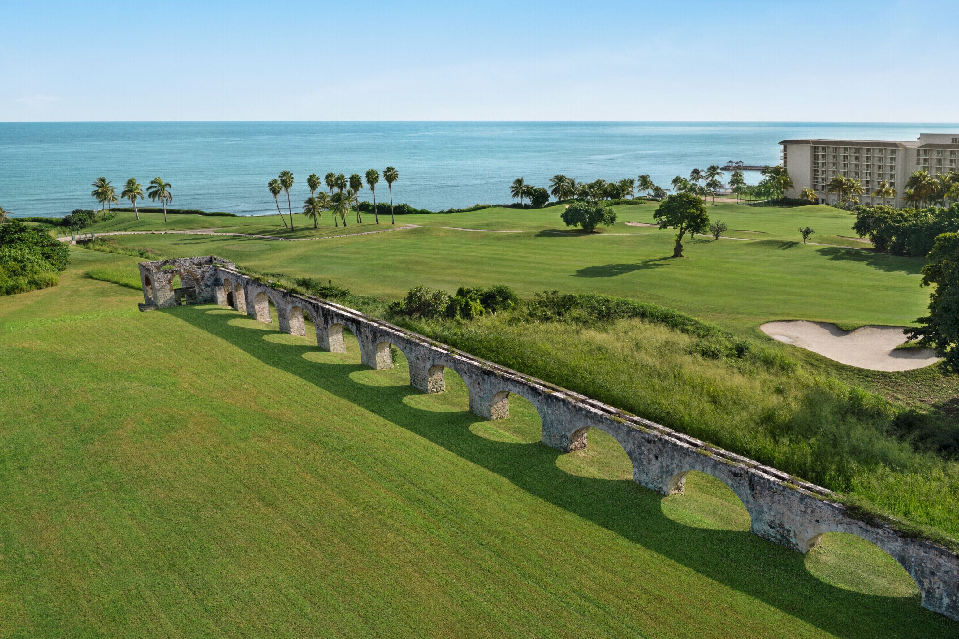 A golf course with a stone bridge, palm trees, and a view of the ocean.