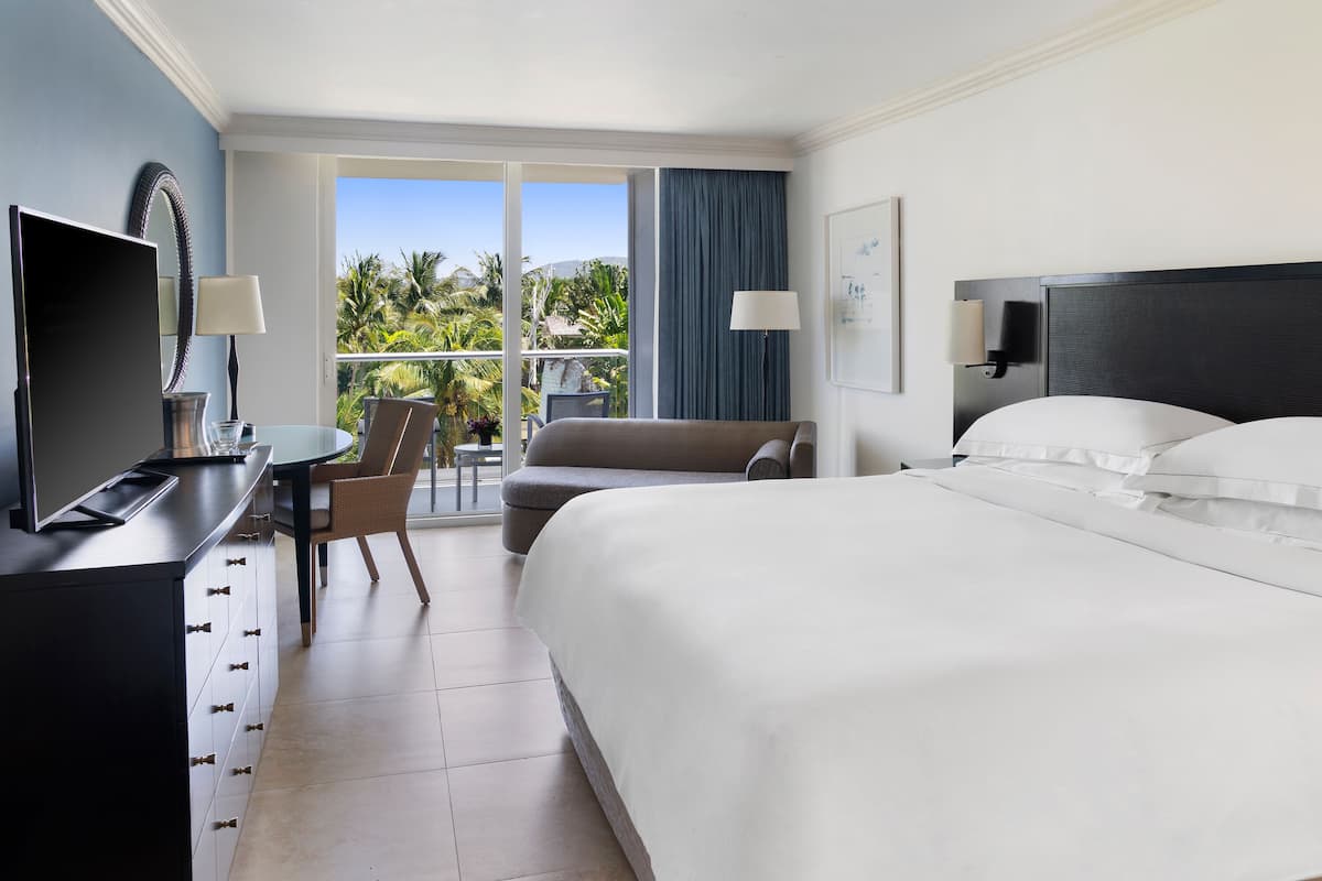 Una camera d'albergo con un letto grande, una scrivania con la televisione, una sedia e vista su palme attraverso la finestra.