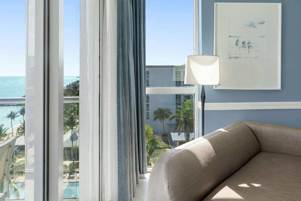 A hotel room with a large window, a sofa, a lamp, and a framed picture on the wall.