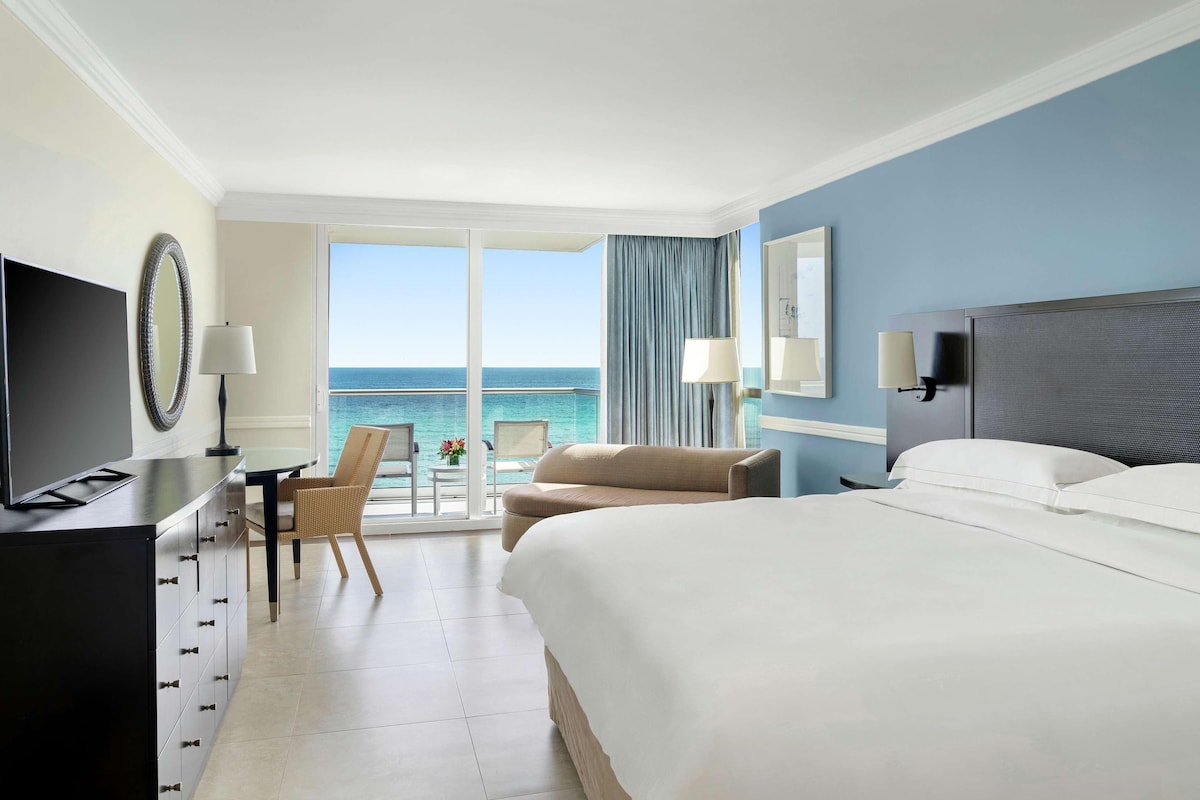 Una camera d'albergo con un letto grande, una scrivania con una sedia, una TV e vista sull'oceano.