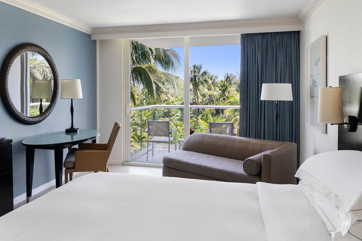 Una camera d'albergo con un letto, un divano, un tavolo con una sedia, una lampada e vista su palme attraverso un'ampia finestra.