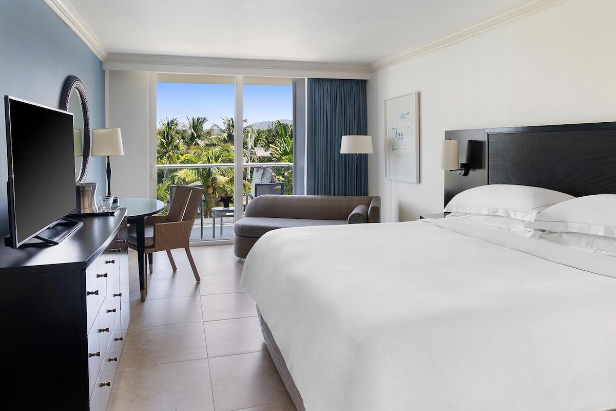 A hotel room with a bed, a desk with a TV, a chair, and a view of palm trees through large windows.