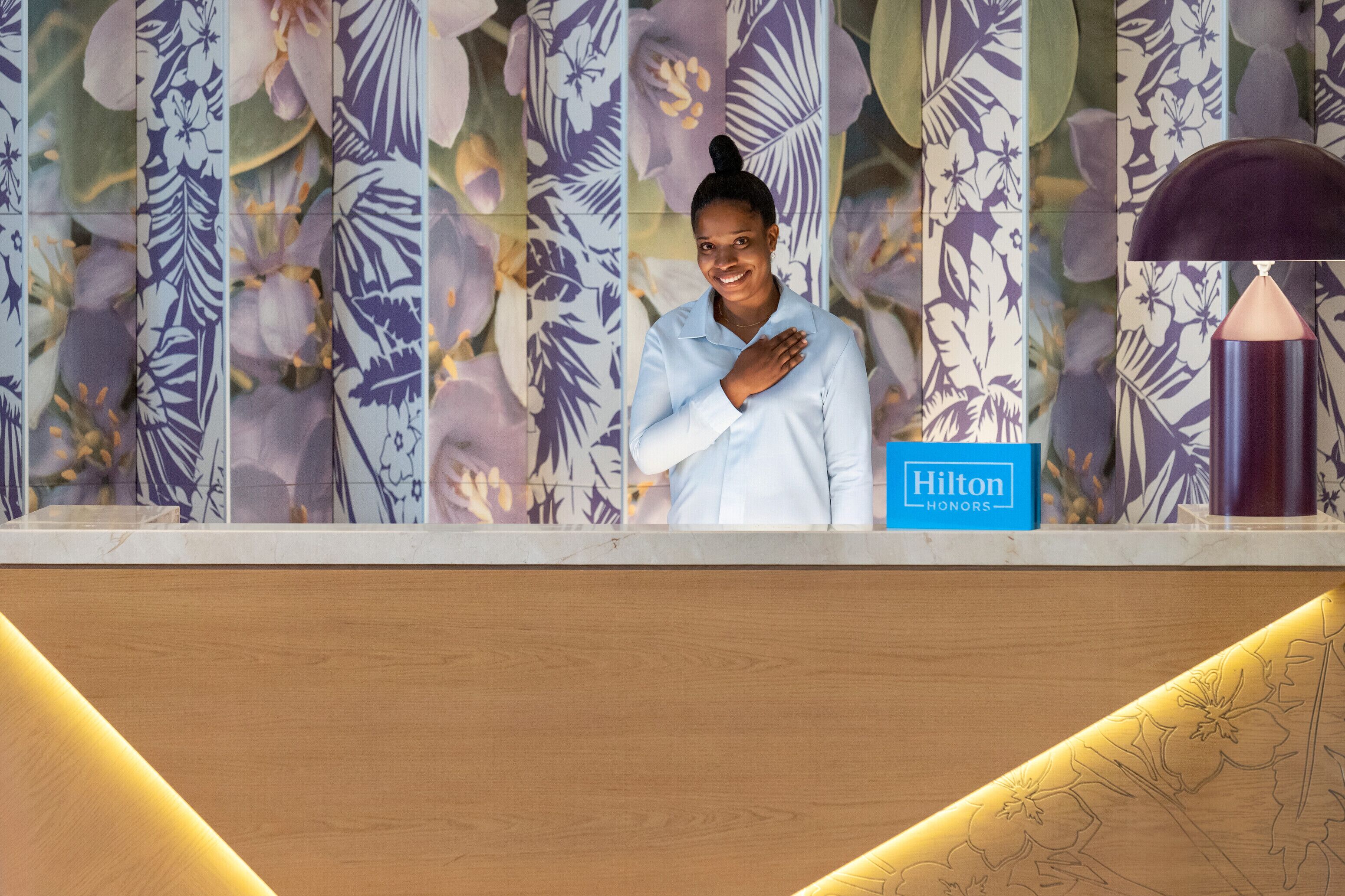 A reception desk with a floral wallpaper background, a blue Hilton Honors sign, and a modern lamp.
