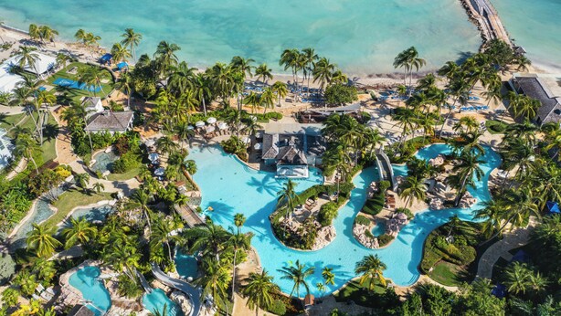 Eine Vogelperspektive zeigt ein Resort mit mehreren Schwimmbecken, Palmen und einem Strandbereich.