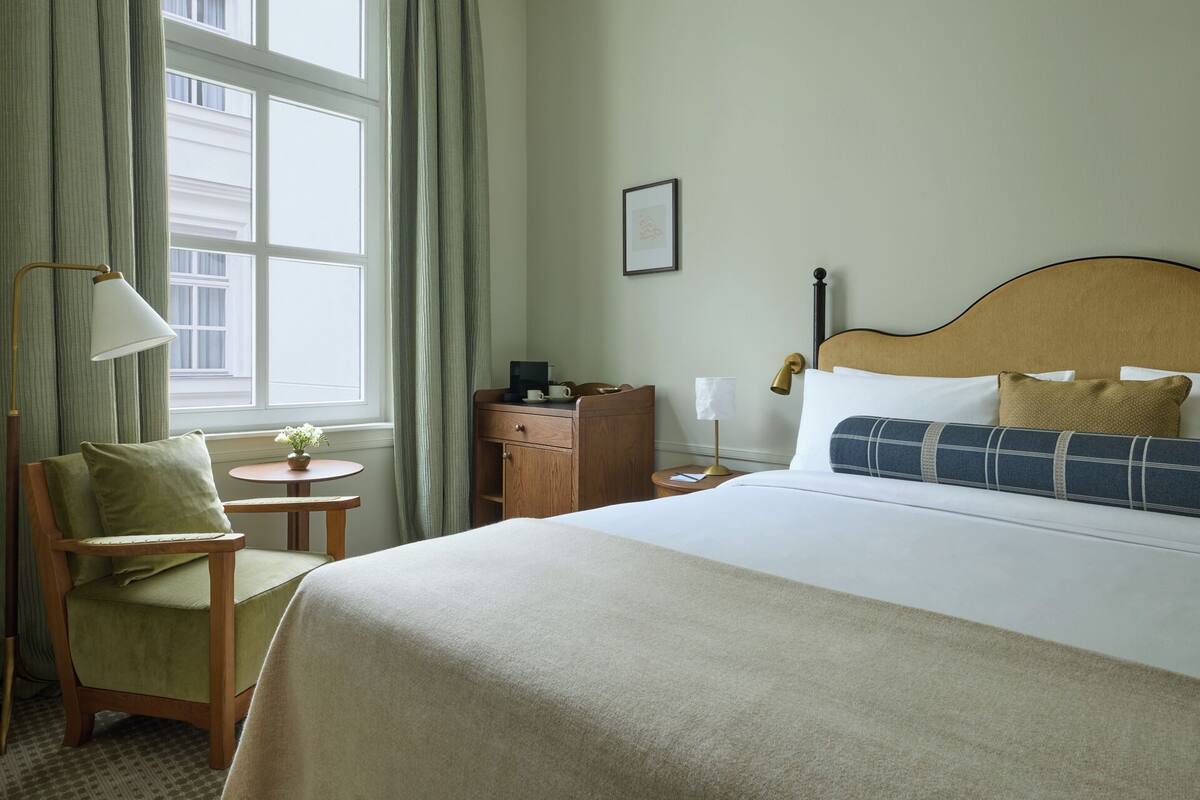 Una habitación de hotel con cama, una silla, mesita de noche, lámpara y ventana con cortinas.