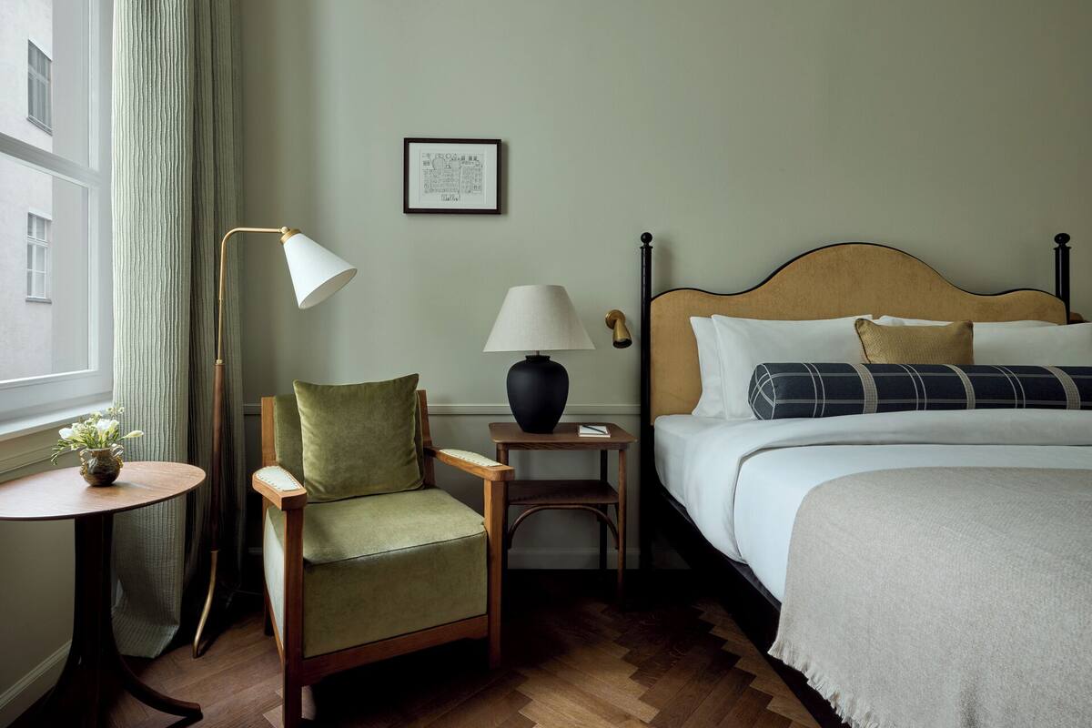 Una habitación de hotel con una cama, una silla, una mesita de noche, una lámpara y un cuadro enmarcado en la pared.