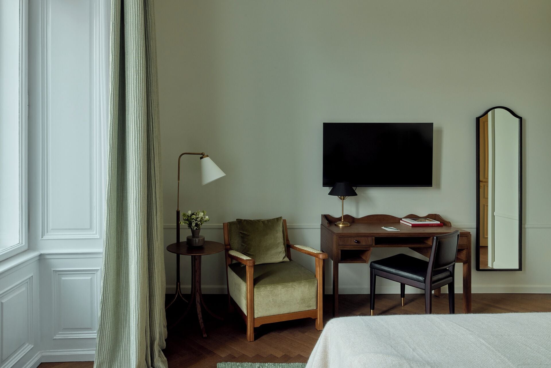 Una habitación de hotel con una cama, un escritorio con una silla, un televisor, un espejo y una mesita con una lámpara y flores.