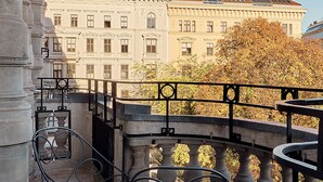 Ein Balkon mit zwei Metallstühlen und einem Tisch, Blick auf ein klassizistisches Gebäude mit zahlreichen Fenstern und einem Baum mit Herbstlaub.