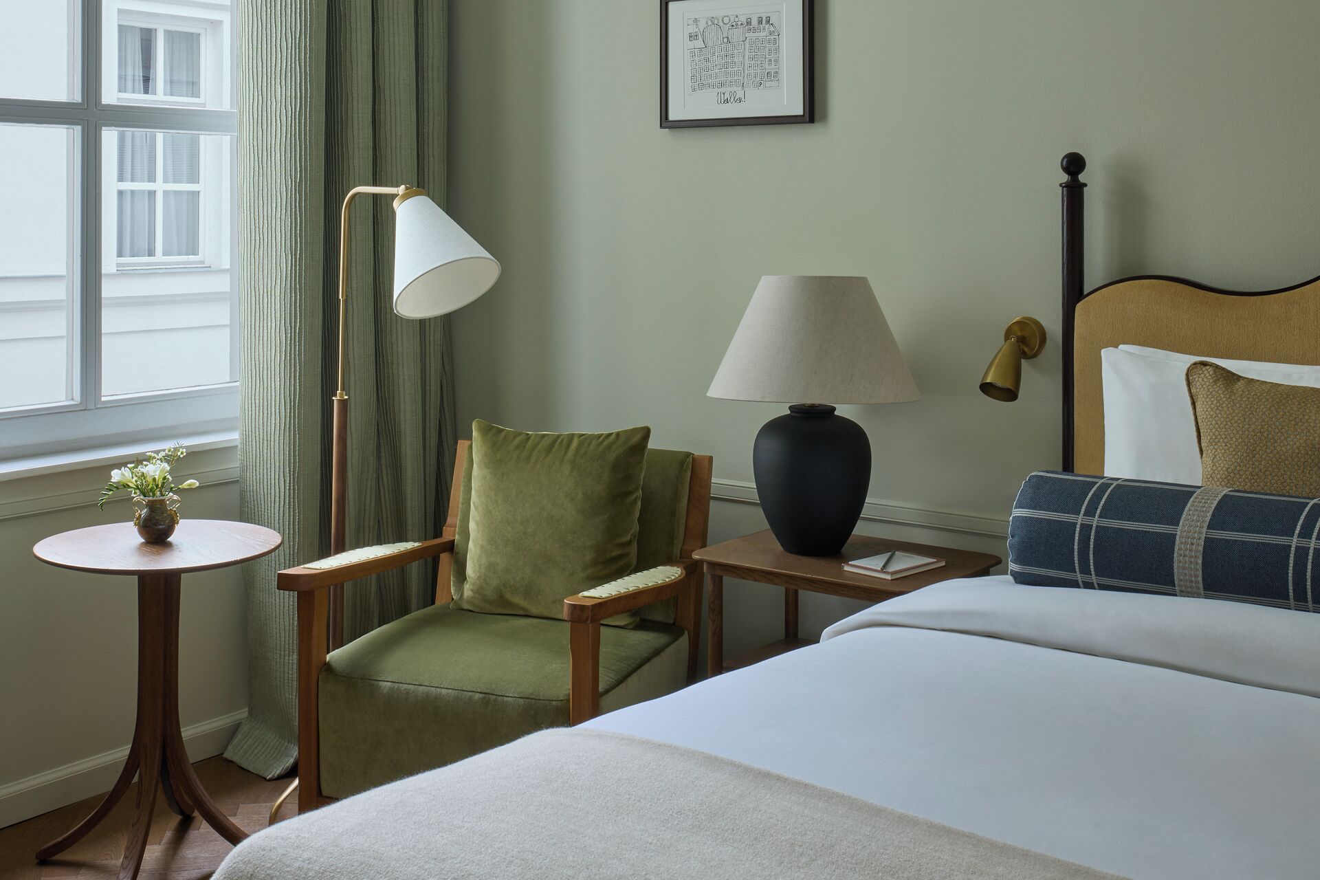 Una habitación de hotel con una cama, un sillón verde, una mesita redonda con un florero, una lámpara de pie y una mesita de noche con una lámpara.