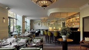 Ein Restaurant mit Barbereich, mehreren Esstischen und Stühlen. Auf einer Seite befinden sich große Fenster, die natürliches Licht hereinlassen.