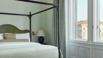 Ein Bett mit Baldachin, dessen Kopfteil grün ist, ein Nachttisch mit Lampe sowie Blick auf eine Stadtkulisse durch ein großes Fenster mit weißem Rahmen.
