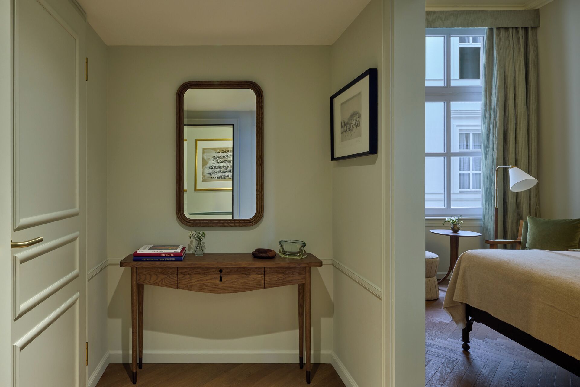 Una camera d'albergo con una scrivania in legno, uno specchio, un quadro incorniciato, un letto, una lampada e una finestra con tende.