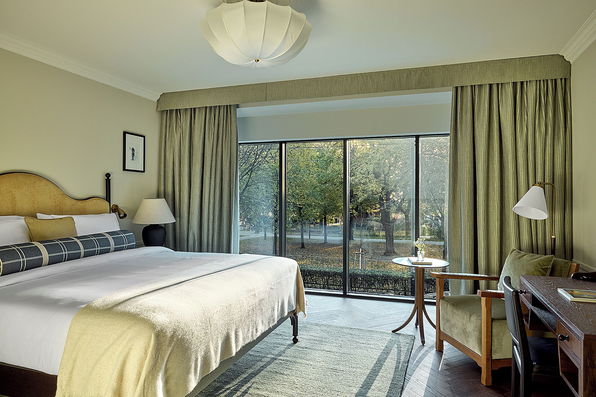 Habitación de hotel con una cama grande, un escritorio, una silla, una lámpara y vistas a árboles por la ventana.