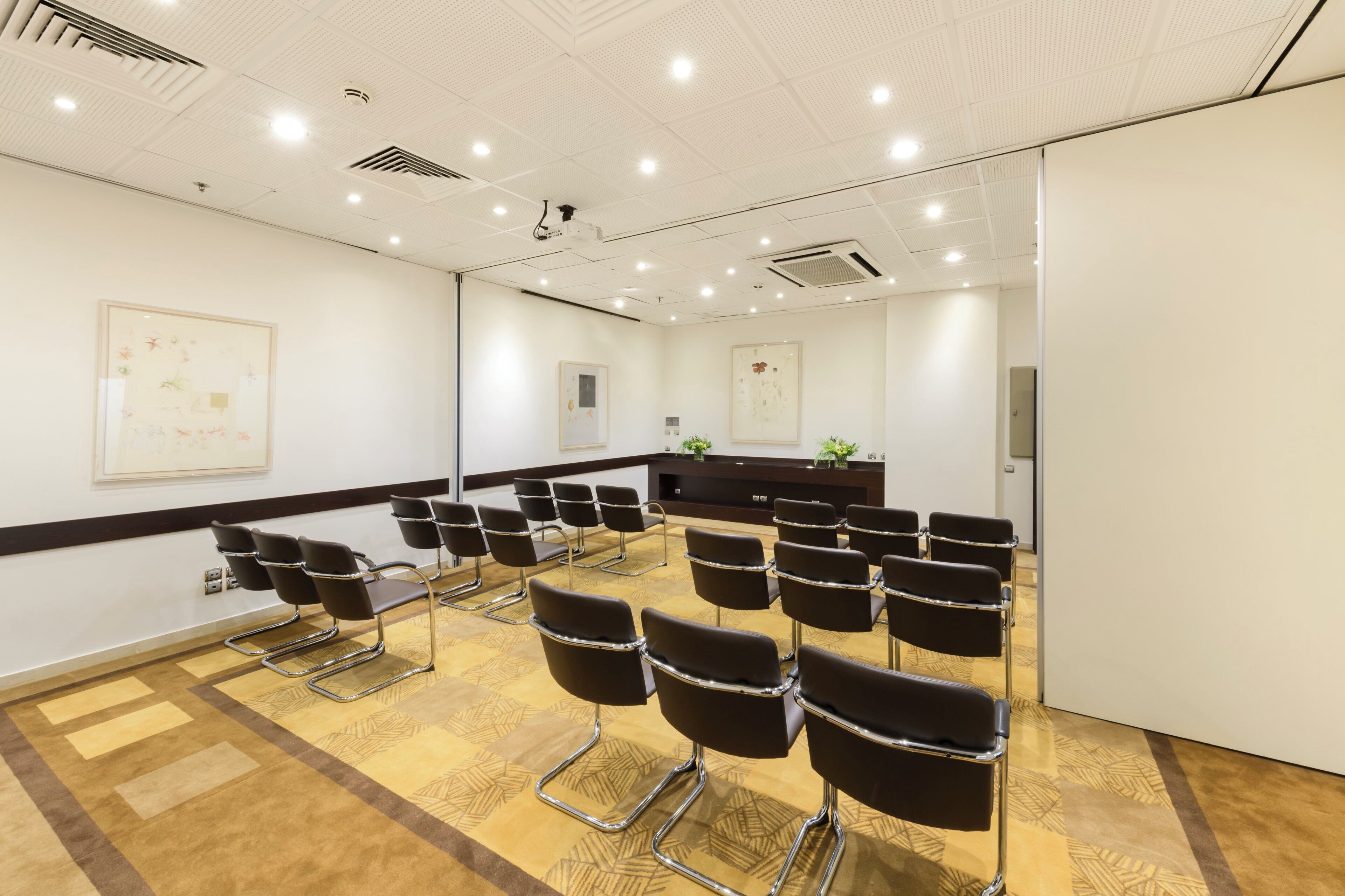 A conference room with rows of chairs, a wooden floor, white walls, and ceiling lights.