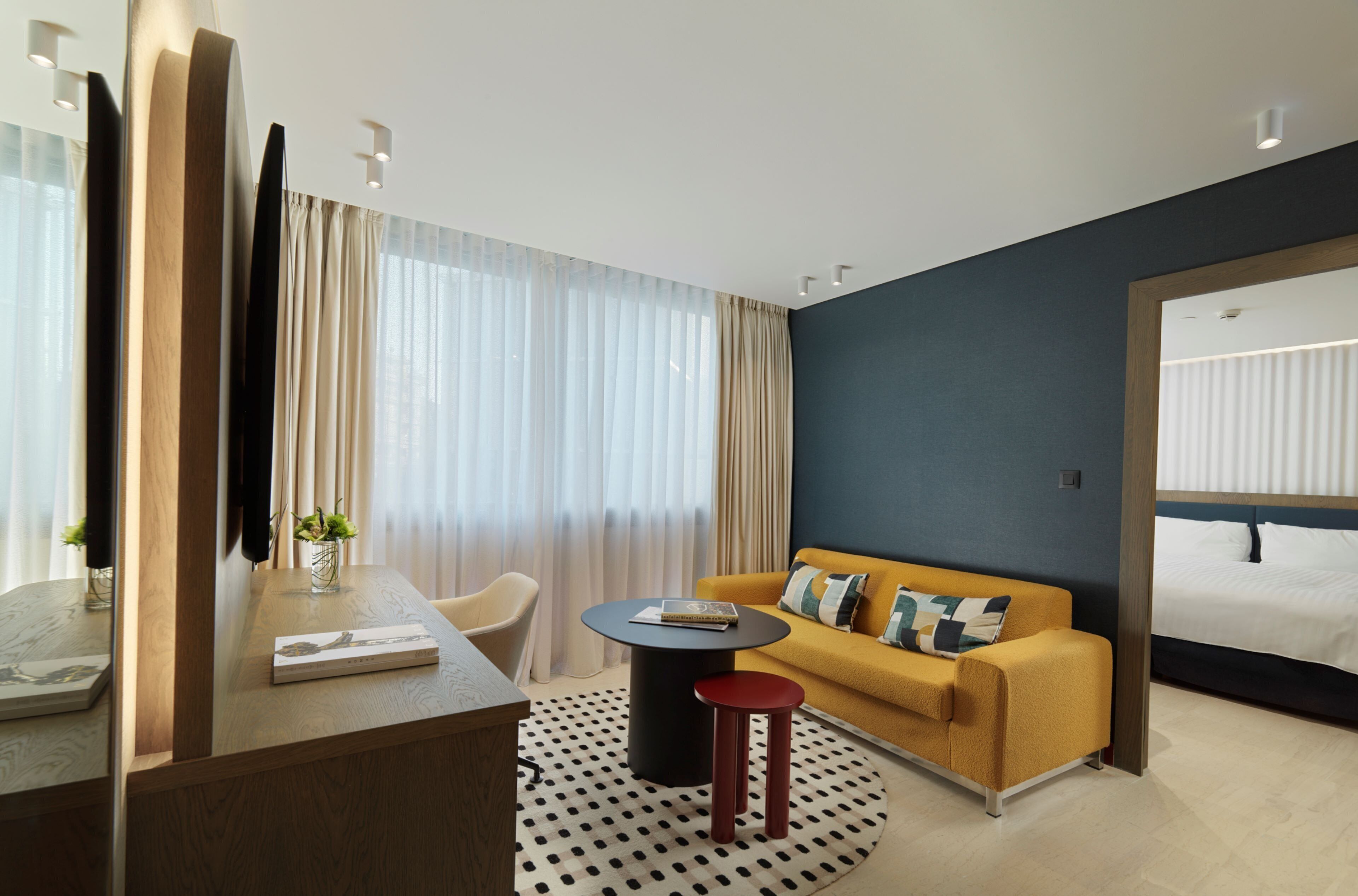 A modern hotel room with a wooden desk, a round black table, a yellow sofa, and a bed with white linens.