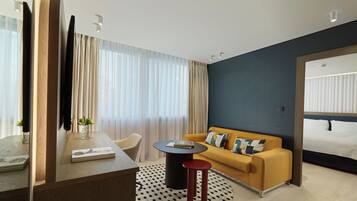 A modern hotel room with a wooden desk, a round black table, a yellow sofa, and a bed with white linens.