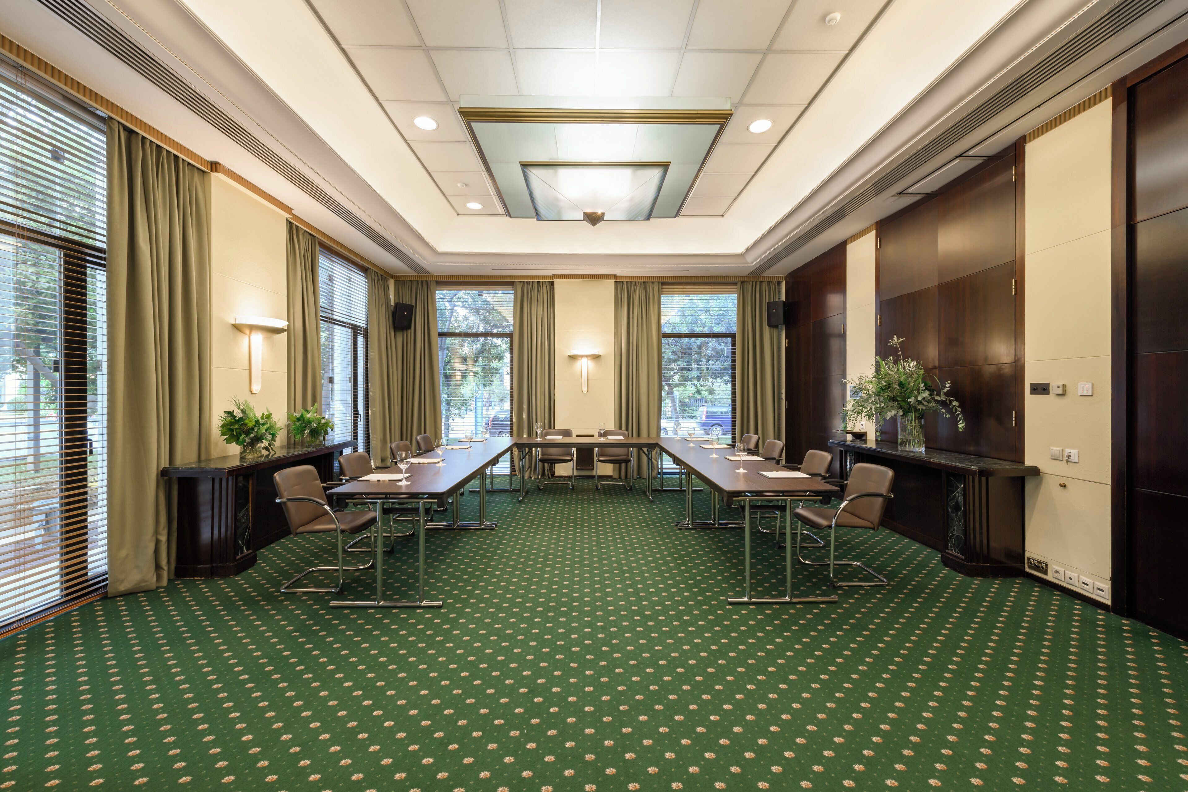 A conference room with a green carpet, long tables, chairs, and a large window with curtains.