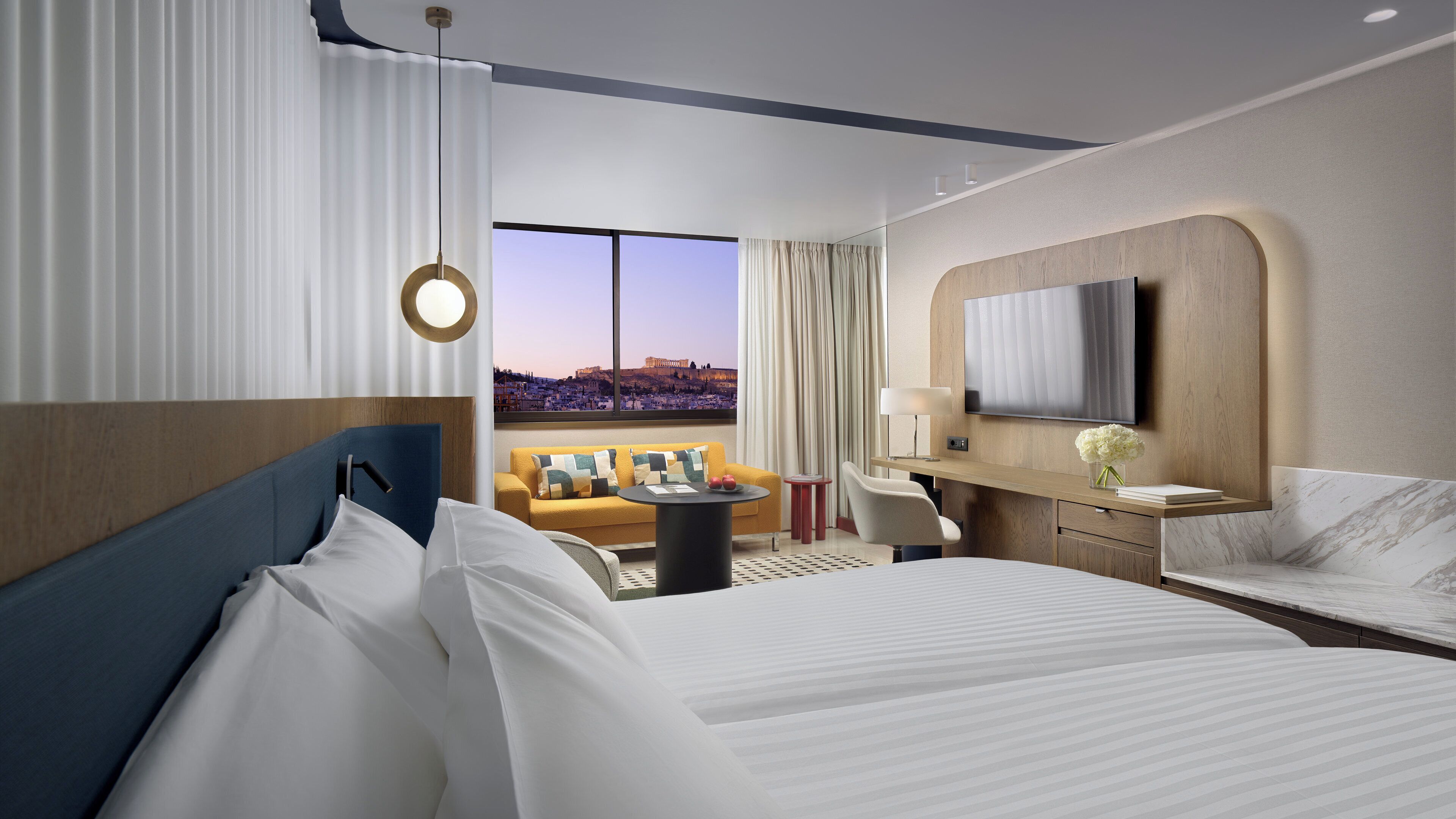 A hotel room with a large bed, a sofa, a TV, and a view of a cityscape.