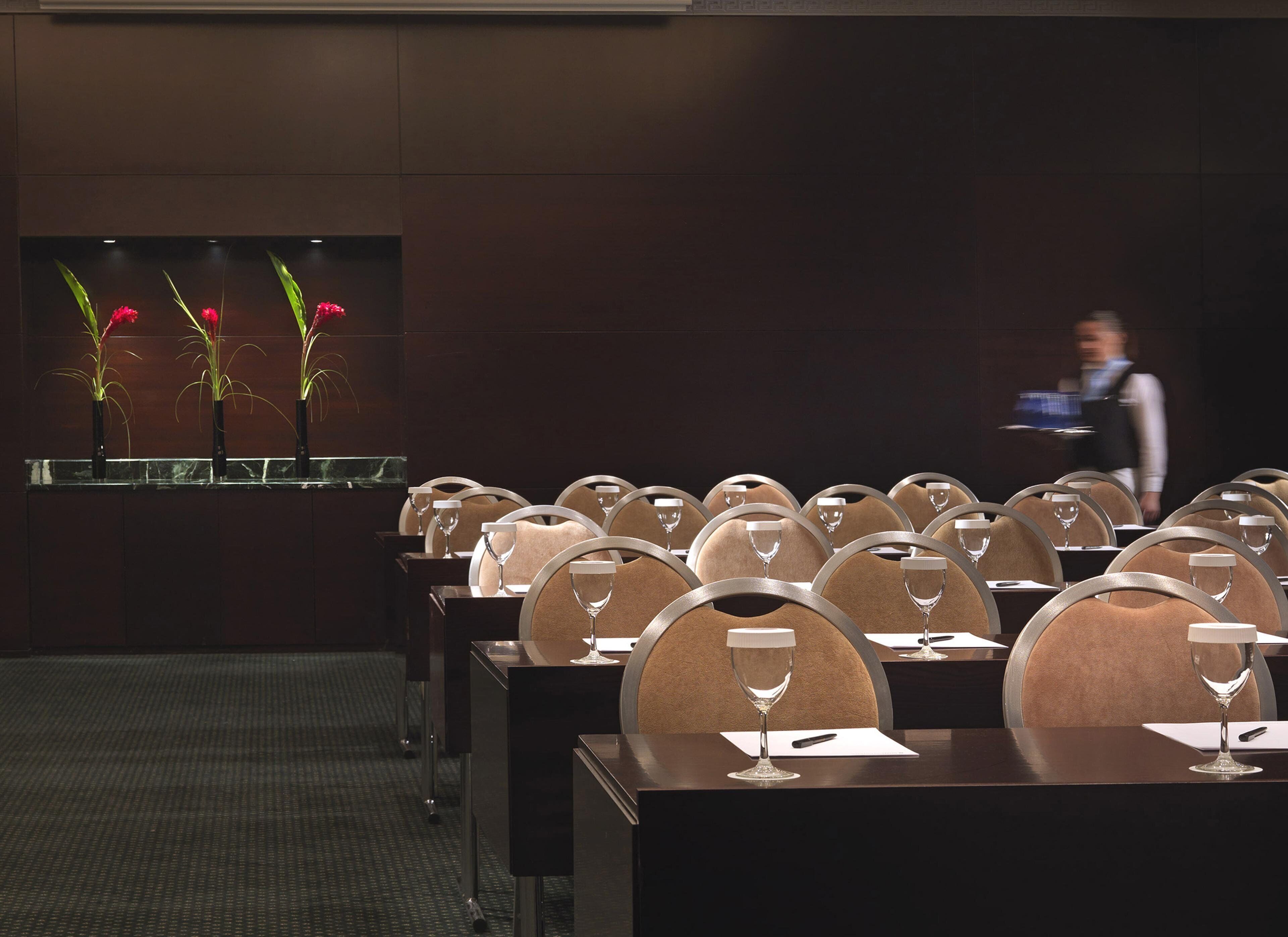 Un salón de conferencias con filas de mesas, cada una con una copa de vino y un bloc de notas. Hay una ventana con plantas a la izquierda.