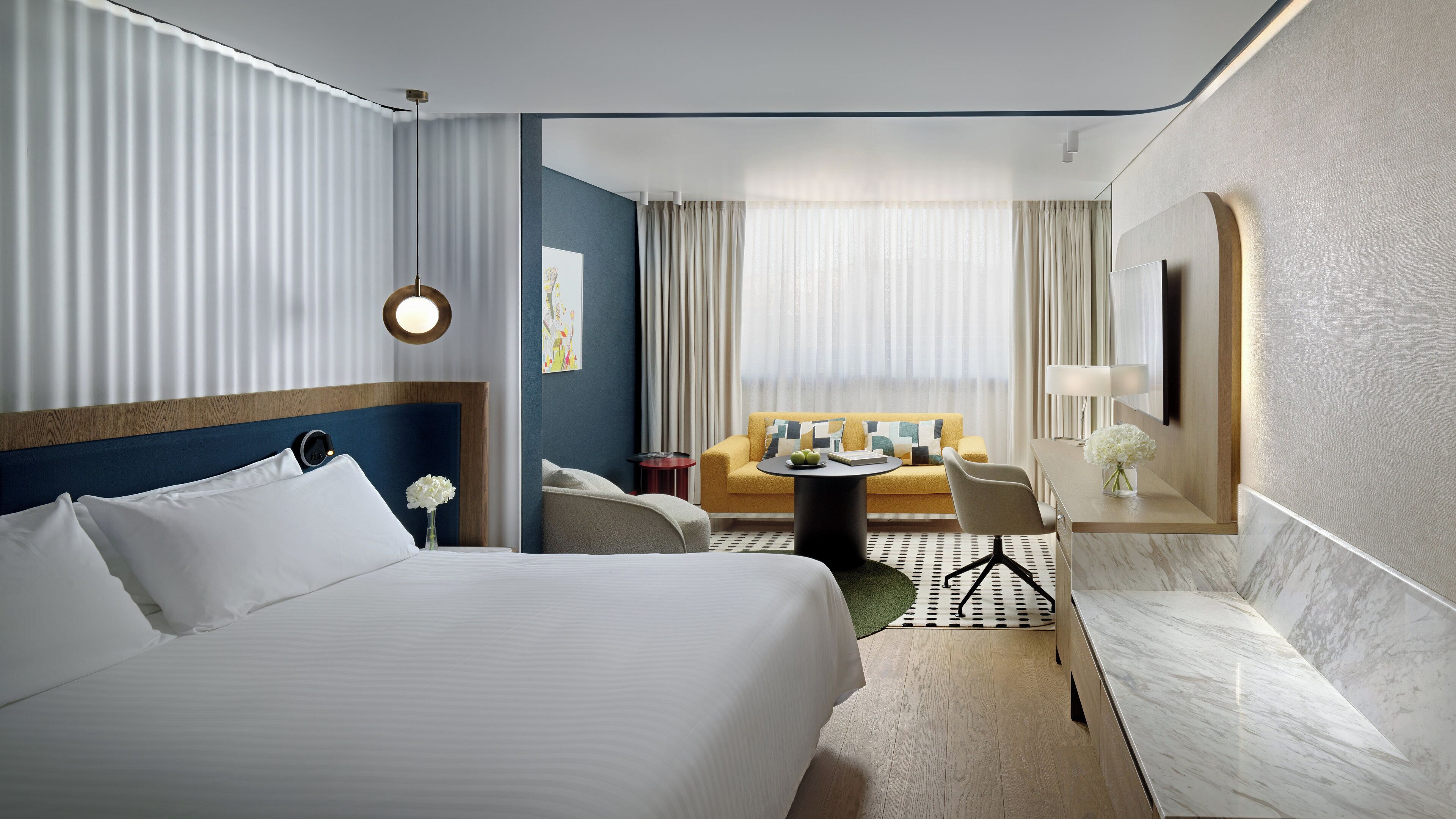A hotel room with a large bed, a sofa, a small round table, and a desk with a lamp.