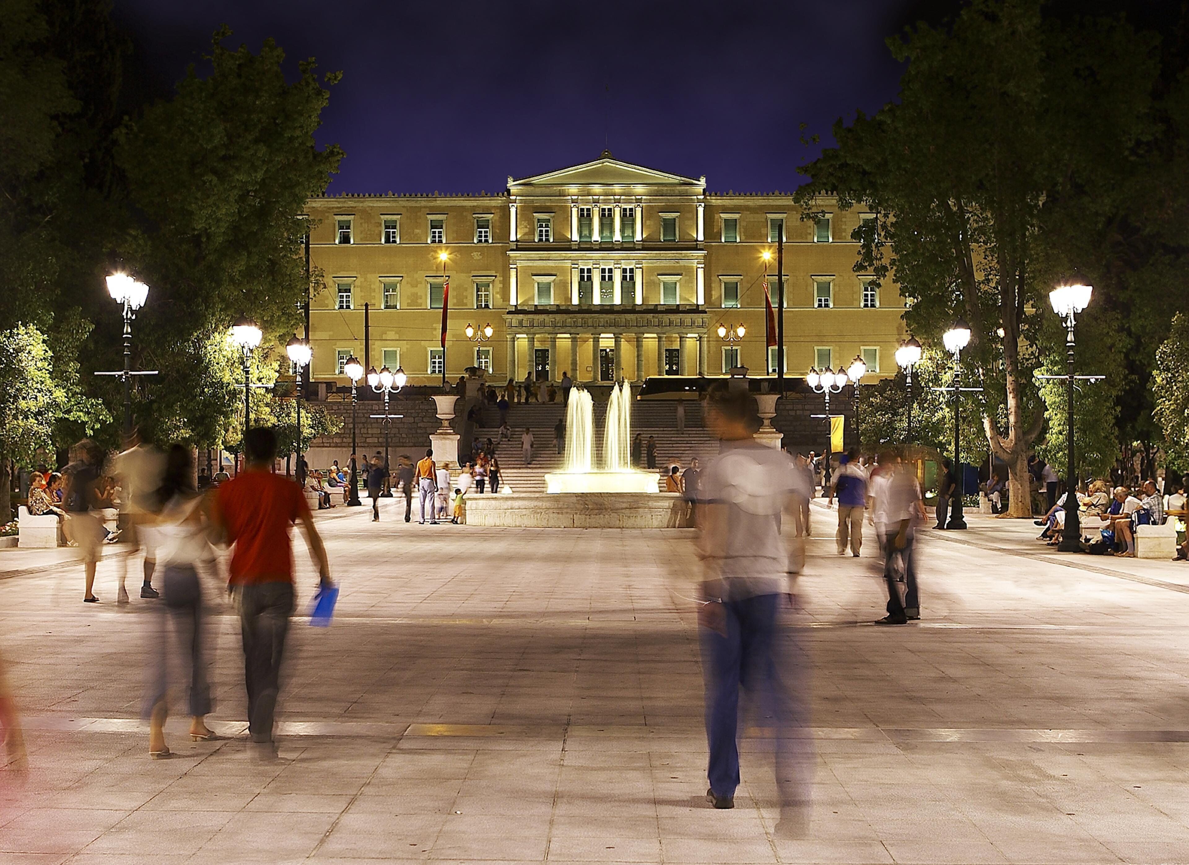 Una plaza pública con un edificio grande al fondo, una fuente y gente caminando.