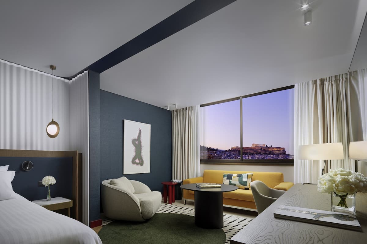 A hotel room with a bed, a sofa, a desk, a chair, a small table, a picture on the wall, and a view of a cityscape through a large window.