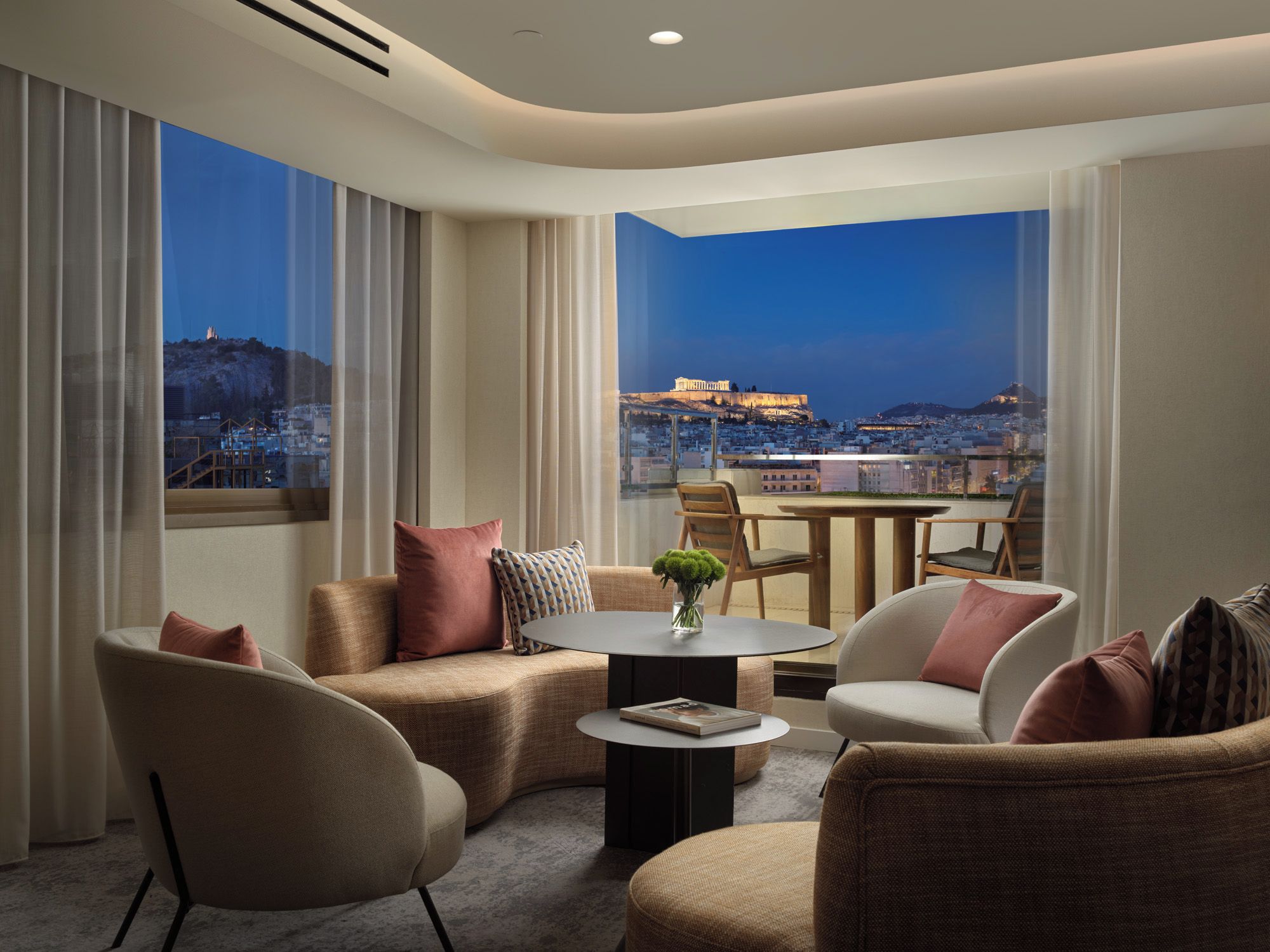 A modern hotel room with a balcony, furnished with sofas, chairs, and a round table.