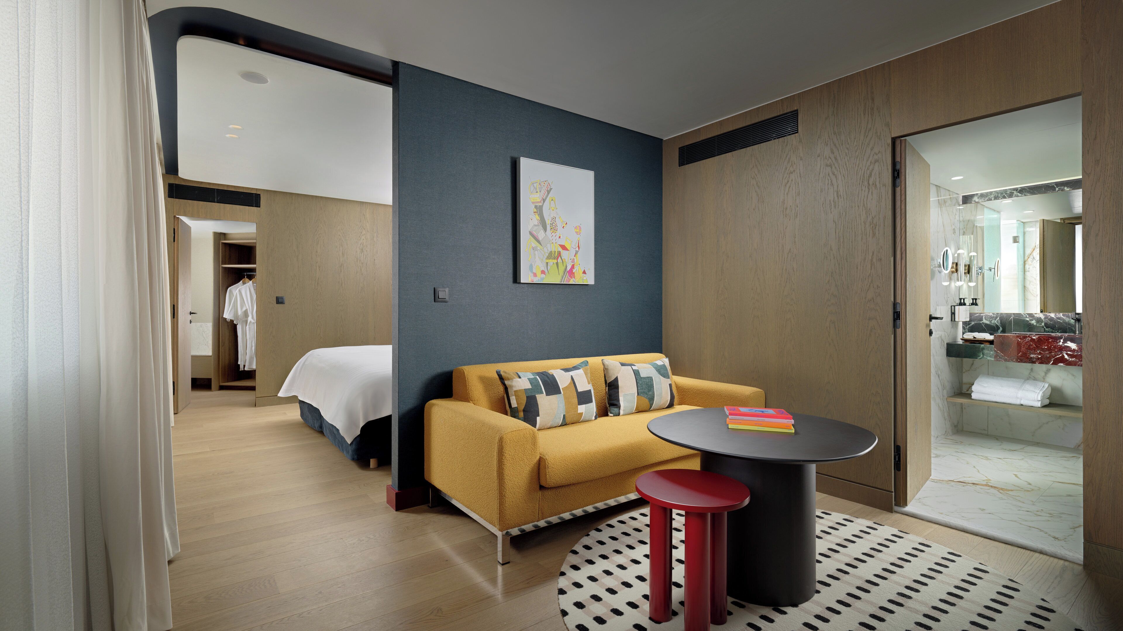 A modern hotel room with a sofa, a small round table, a bed, and a bathroom visible through an open door.