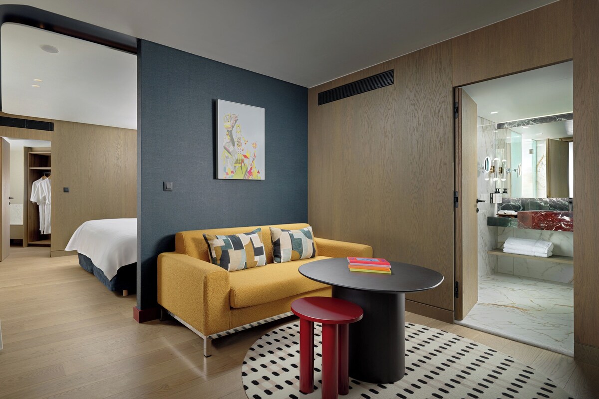 A modern hotel room with a sofa, a small round table, a bed, and a bathroom visible through an open door.