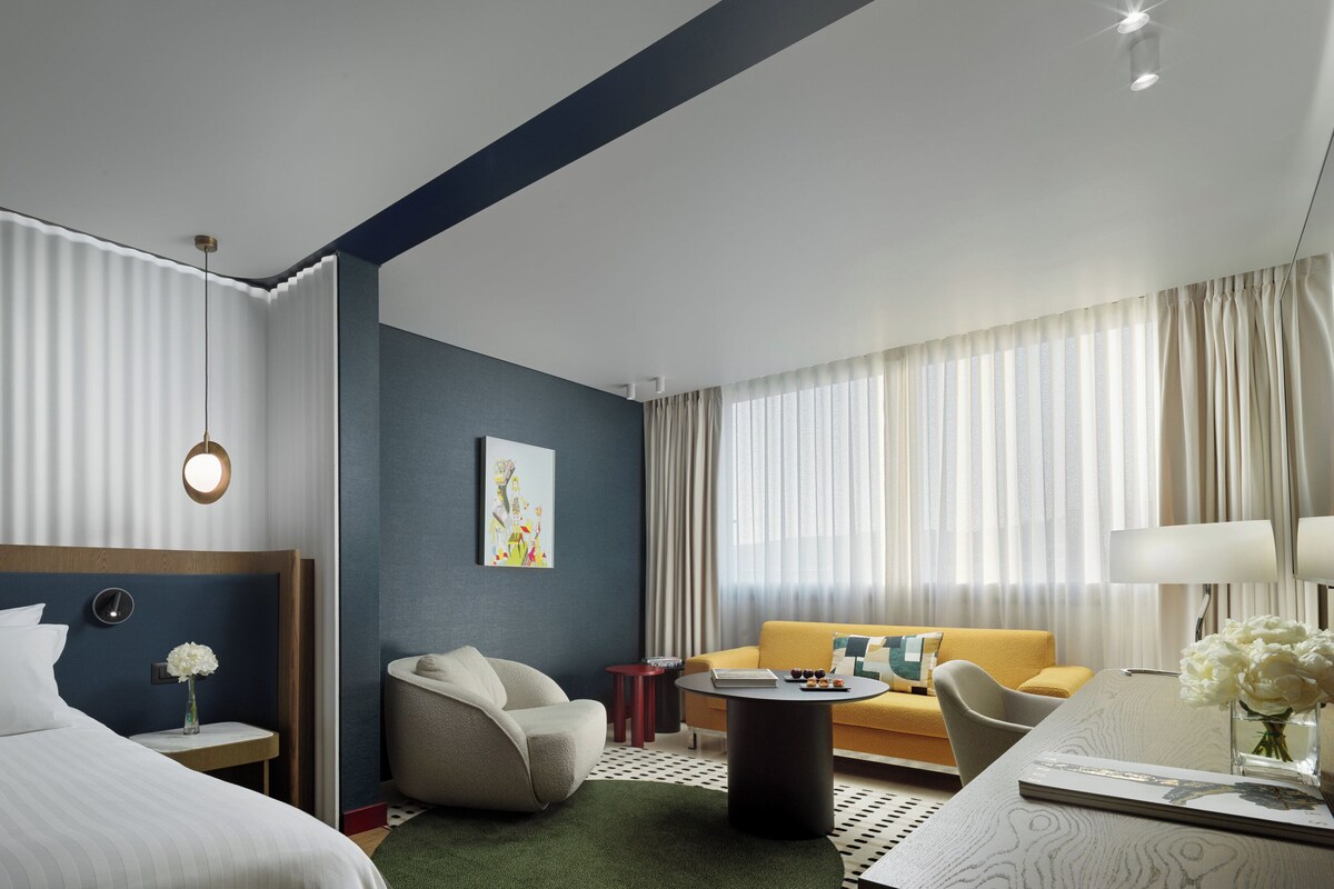 A modern hotel room with a bed, a sofa, a coffee table, and a desk with a lamp.