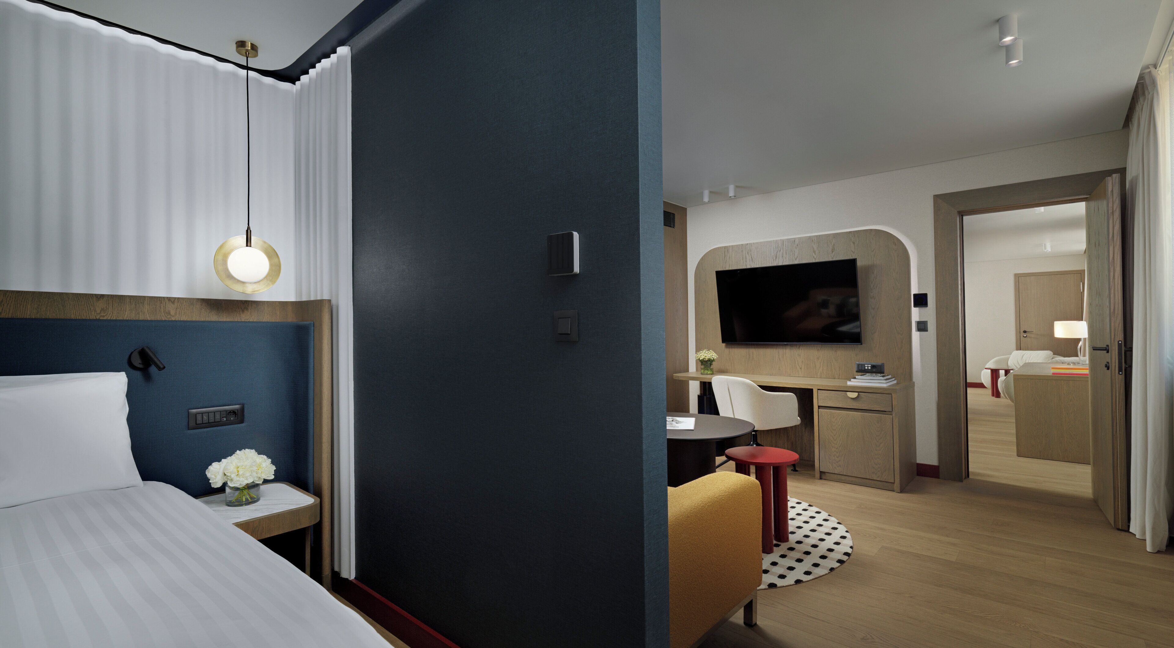 Habitación de hotel moderna con cama, mesita de noche y escritorio con silla. Hay un televisor montado en la pared y una puerta que da a otra habitación.